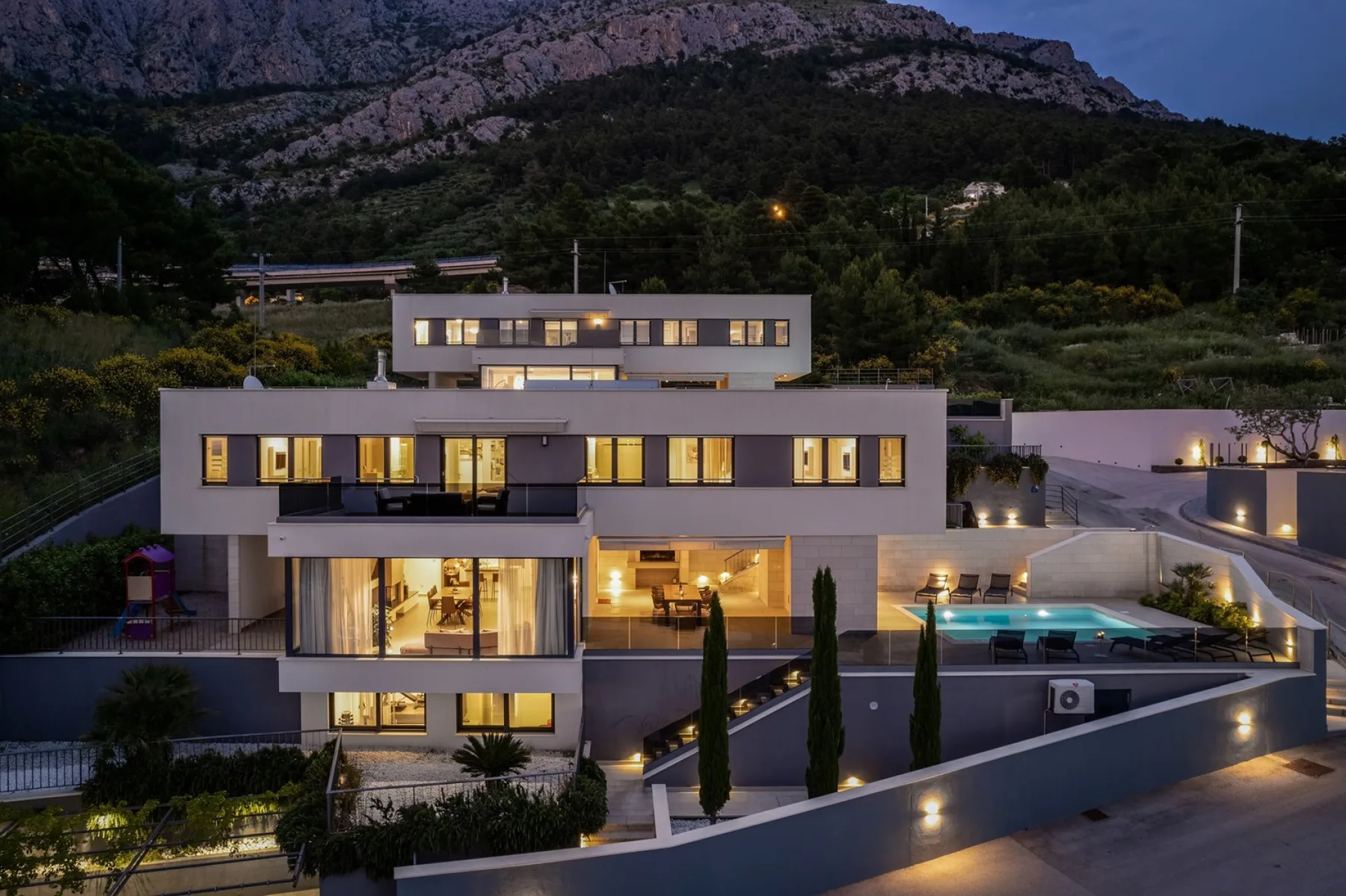 A modern hillside house overlooks an illuminated outdoor pool at dusk.
