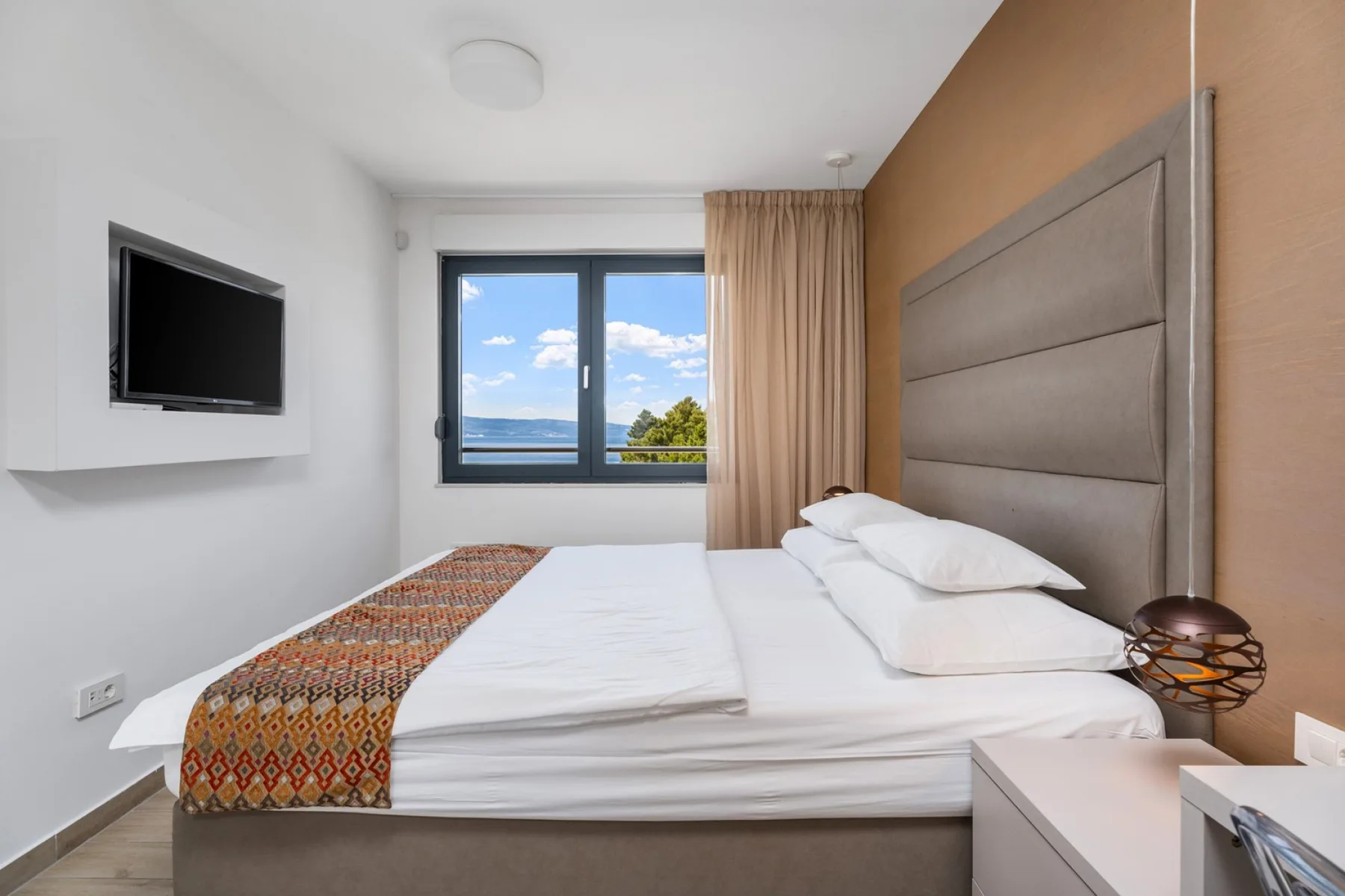 A bedroom with a neatly made bed, wall-mounted television, and a window overlooking the sea.