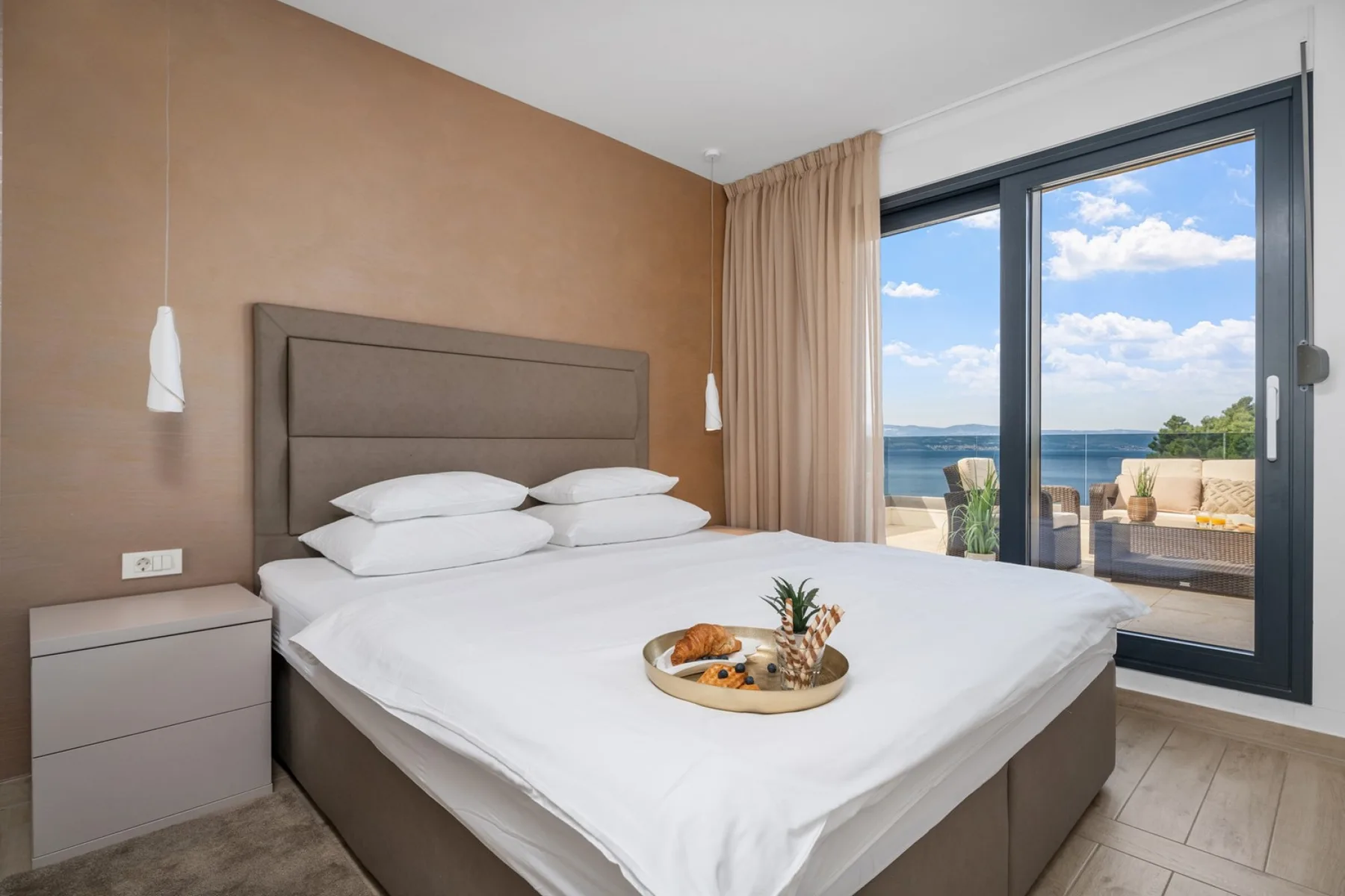 A bedroom with a neatly made bed beside large glass doors overlooking the sea.