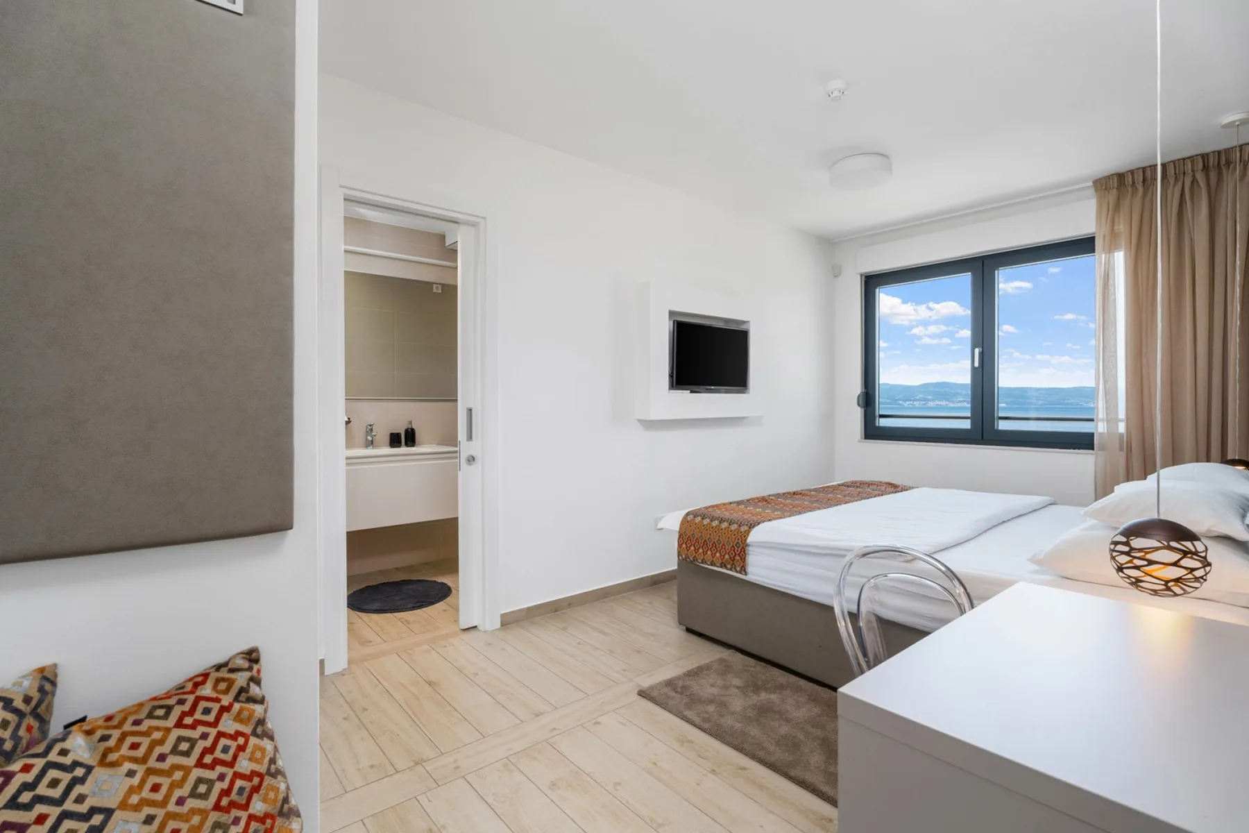 A bright bedroom with a bed, wall-mounted television, and large windows.