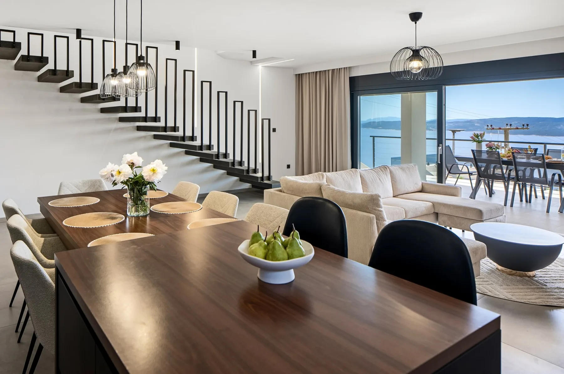 An open-plan living and dining room features a long wooden table, a staircase, and wide windows overlooking the water.