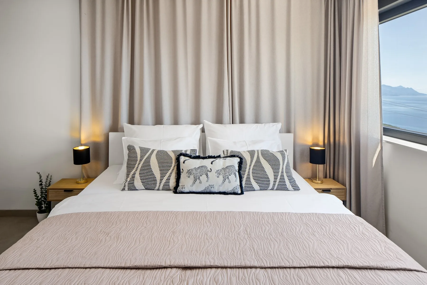 A neatly made bed with patterned cushions sits between bedside lamps, with curtains and a sea view through the window.