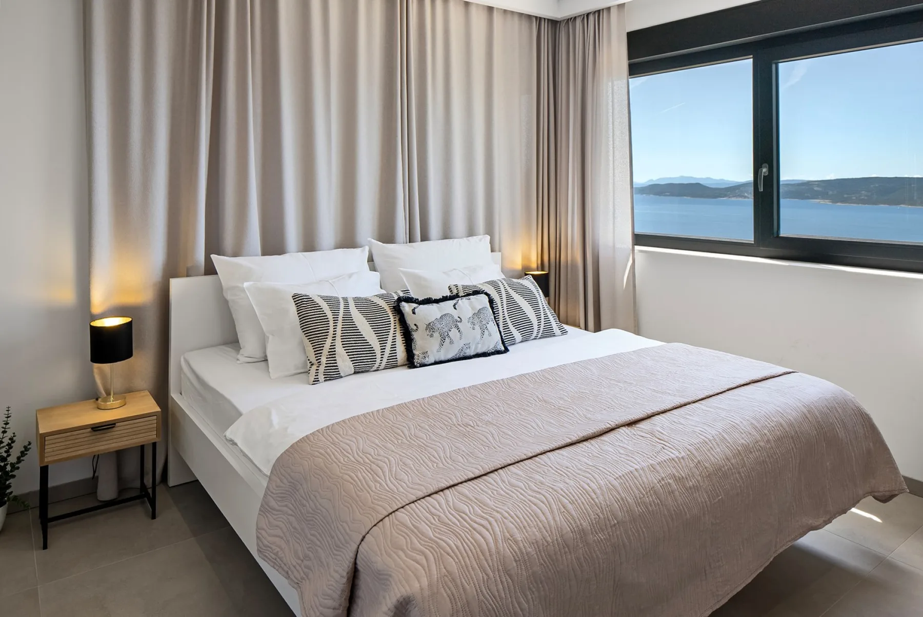 A neatly made bed with patterned cushions sits beside a large window overlooking the water.