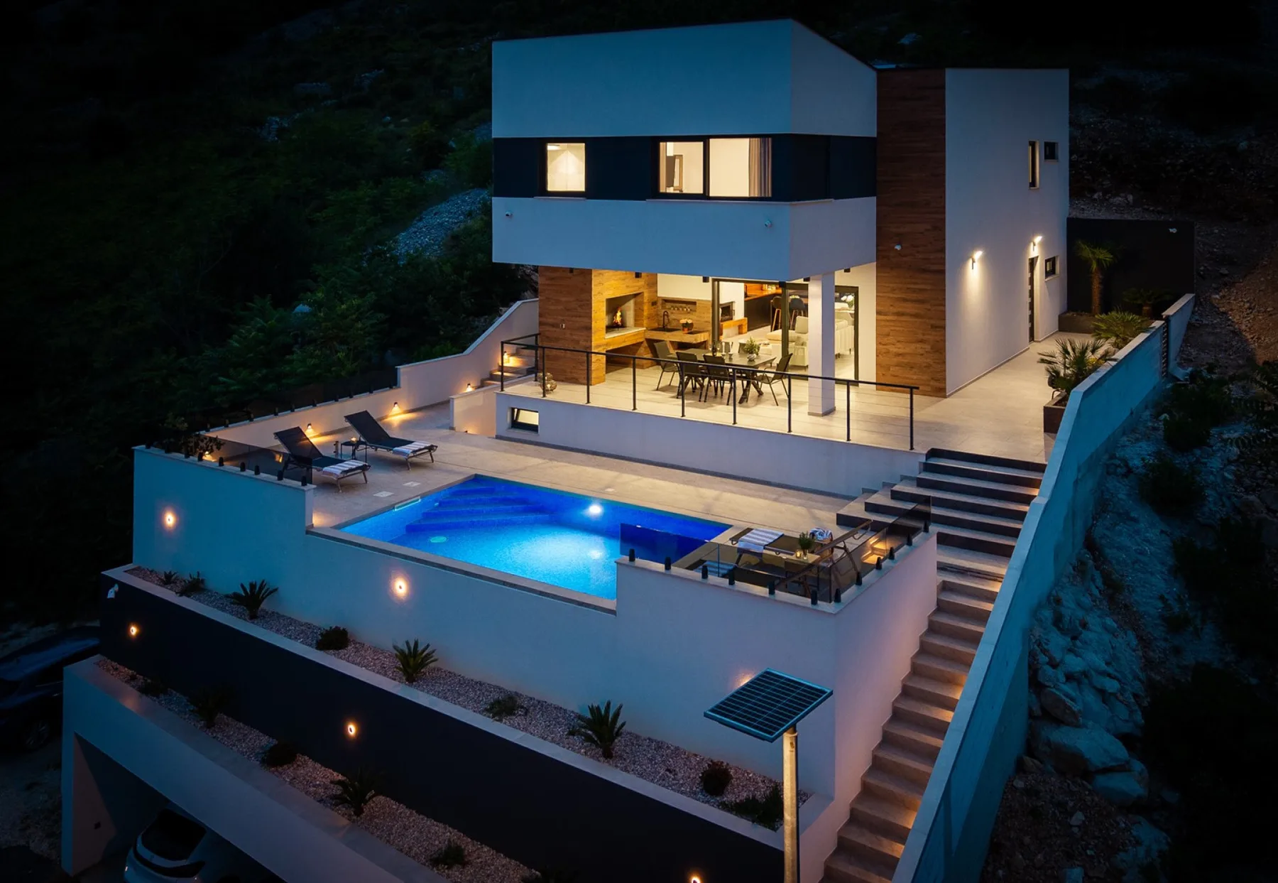 A modern two-story house overlooks a lit swimming pool, with outdoor dining and lounge areas.