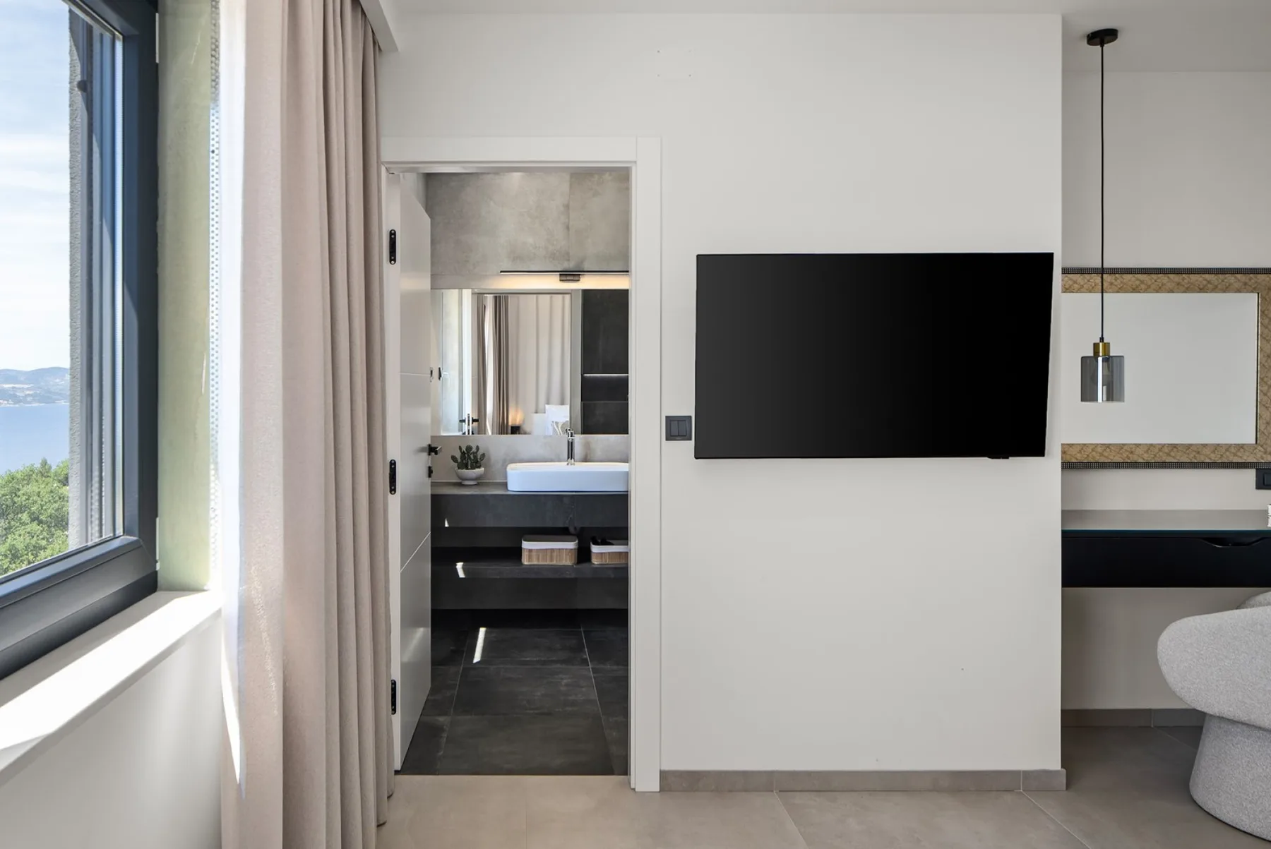A modern bedroom area with a wall-mounted television, floor-to-ceiling window, and an open doorway to the bathroom.