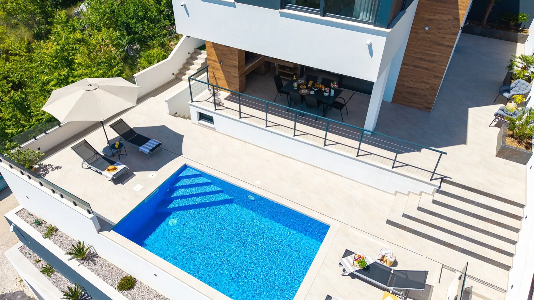 A modern house opens onto a bright patio with a blue swimming pool, lounge chairs, and a shaded dining area.