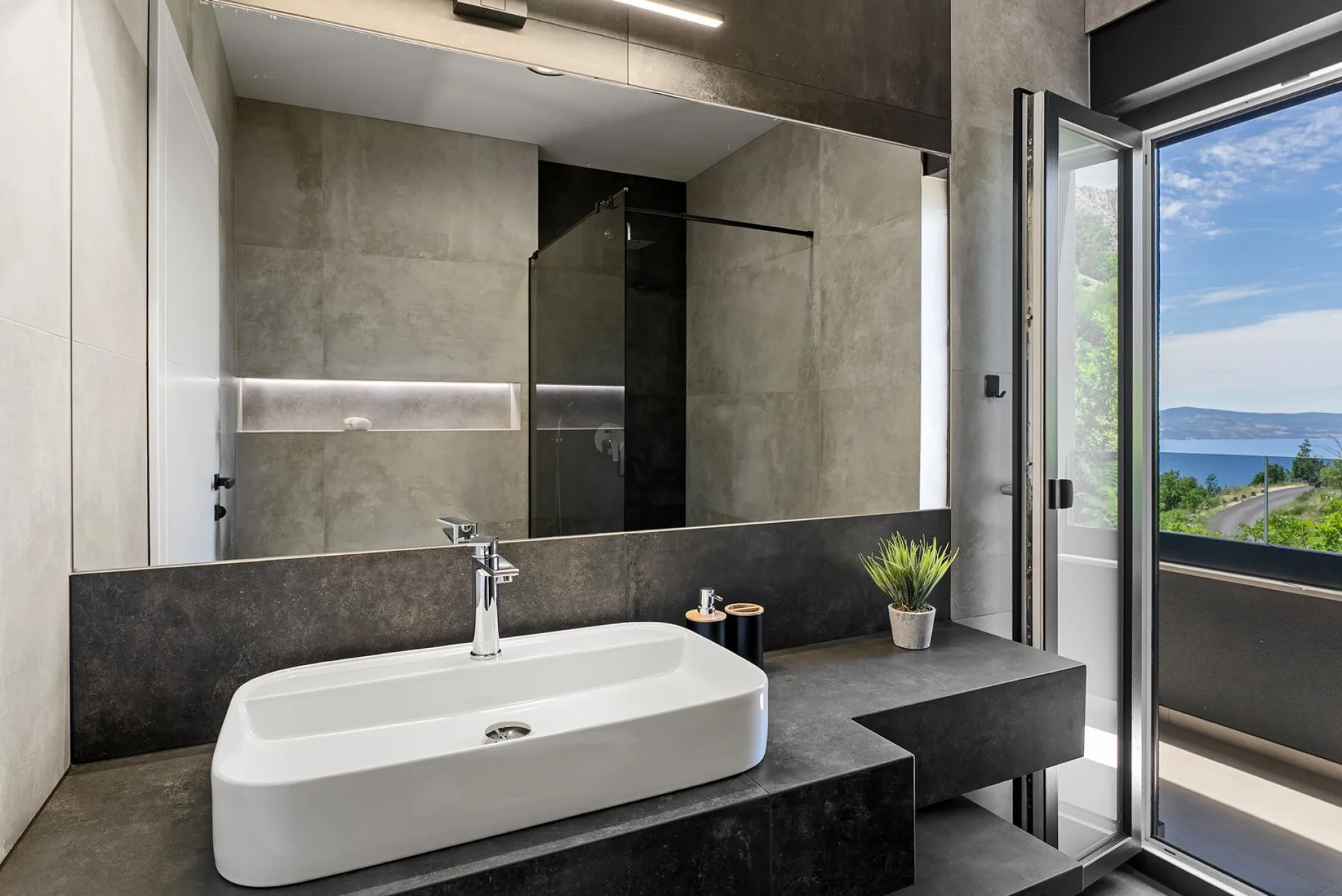 A modern bathroom has a white vessel sink on a dark counter, with a glass shower and open balcony door.