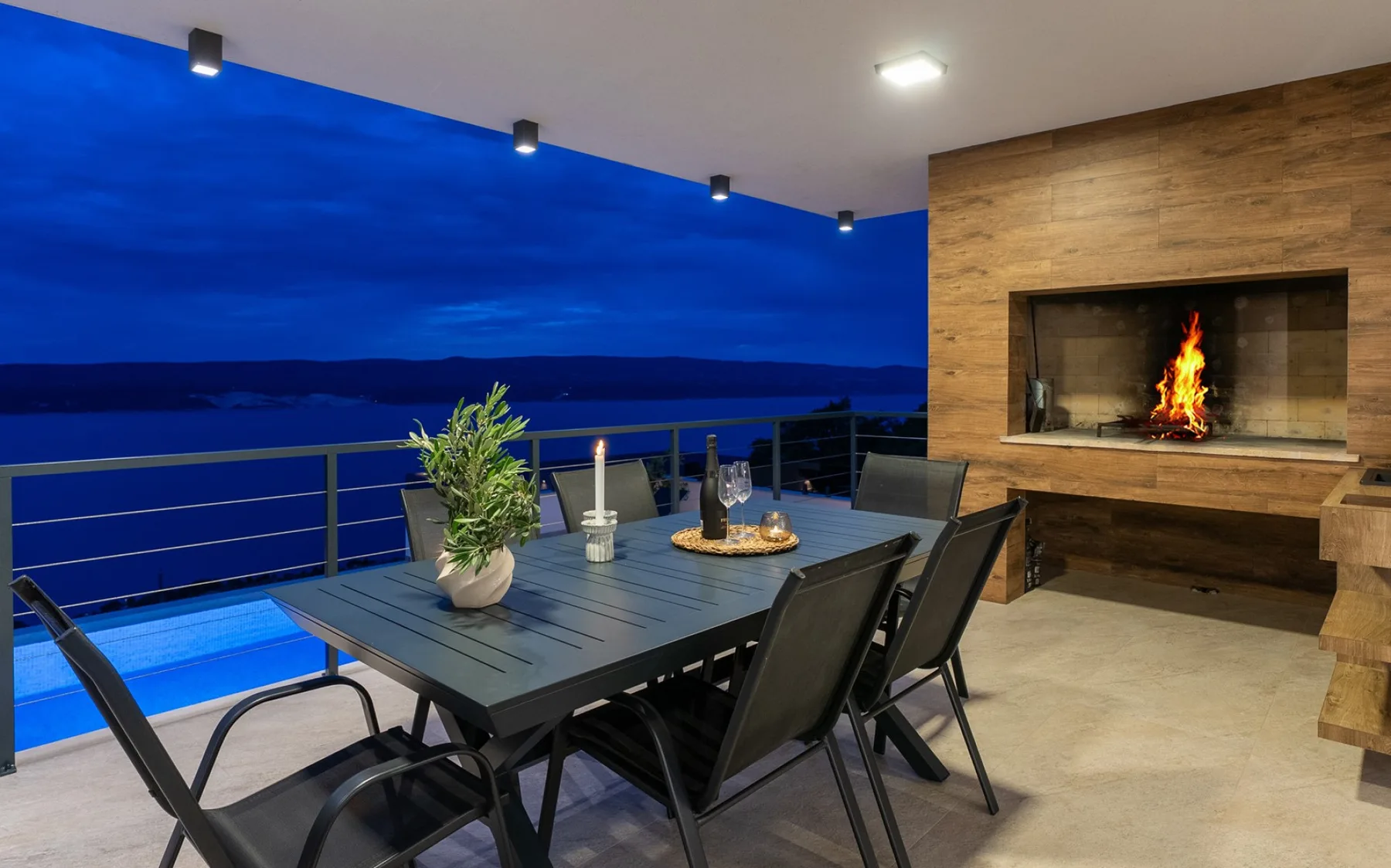 An outdoor dining terrace overlooks the water, with a table, chairs, and a built-in fireplace.