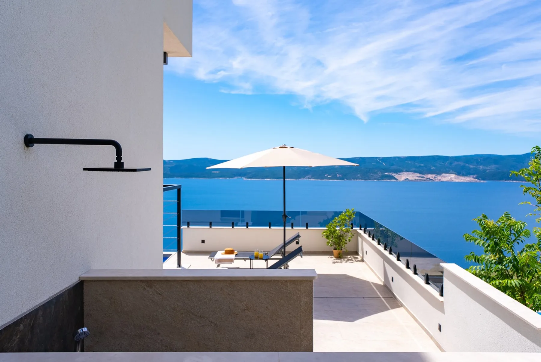 A sunlit terrace overlooks blue water, with a white umbrella, lounge chairs, and glass railings.