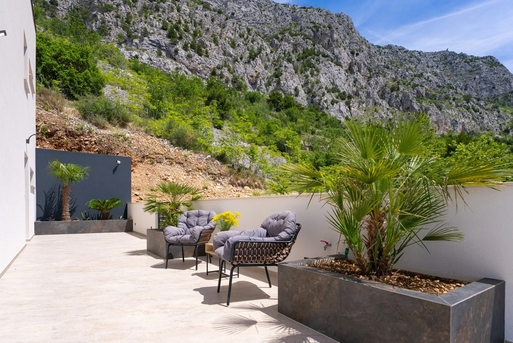 A sunlit outdoor patio has cushioned lounge chairs, large planters, and a rocky hillside beyond the wall.