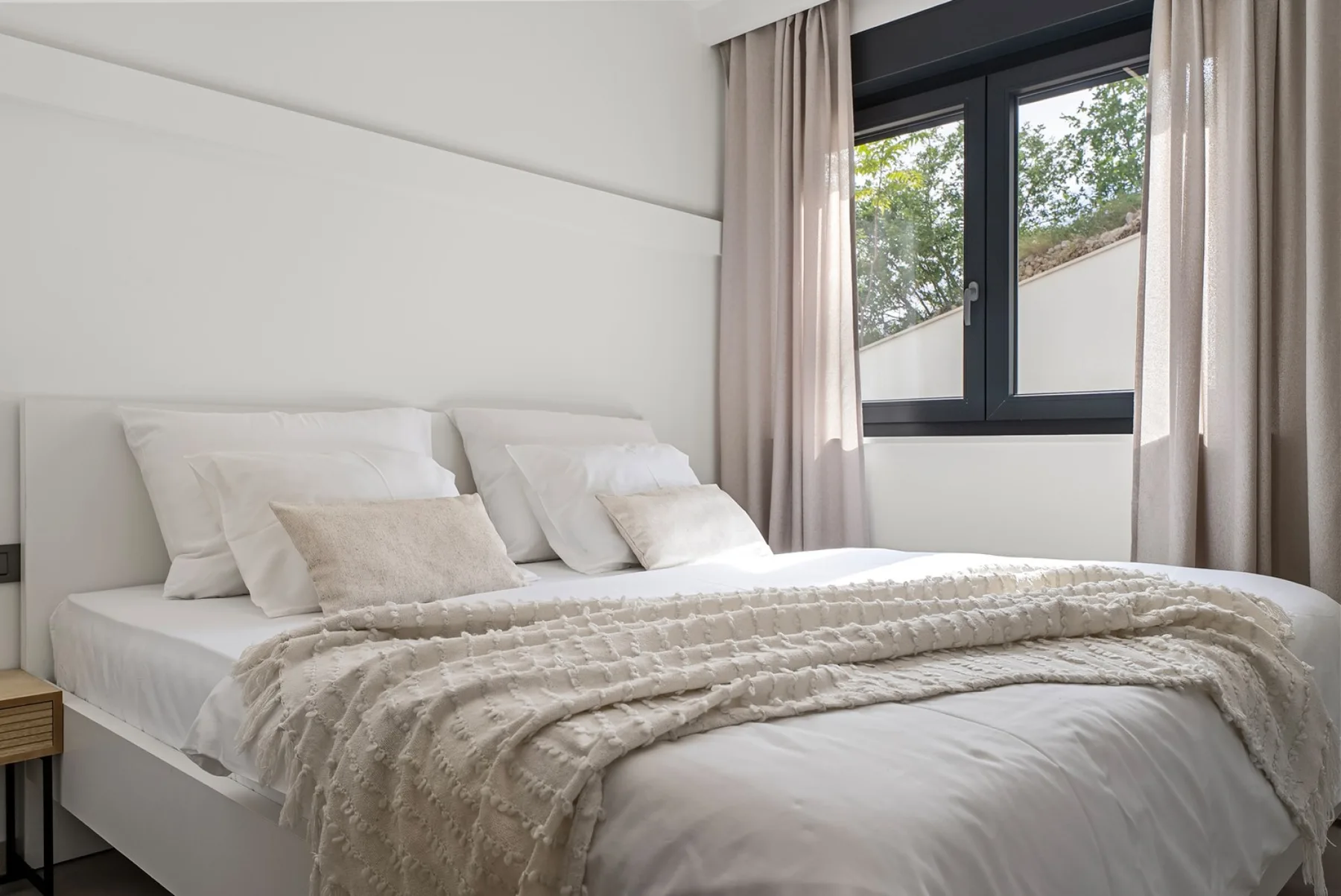 A neatly made bedroom features a white bed with layered pillows, a textured throw, and a large curtained window.