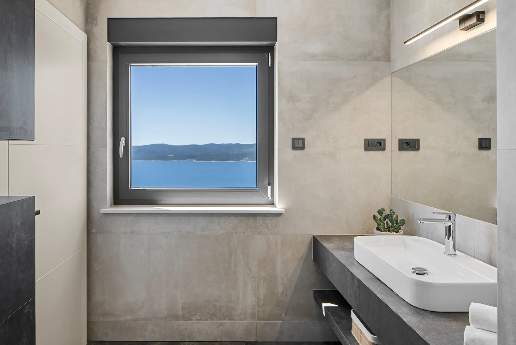 A modern bathroom with a wide window overlooking blue water and hills, beside a white vessel sink.