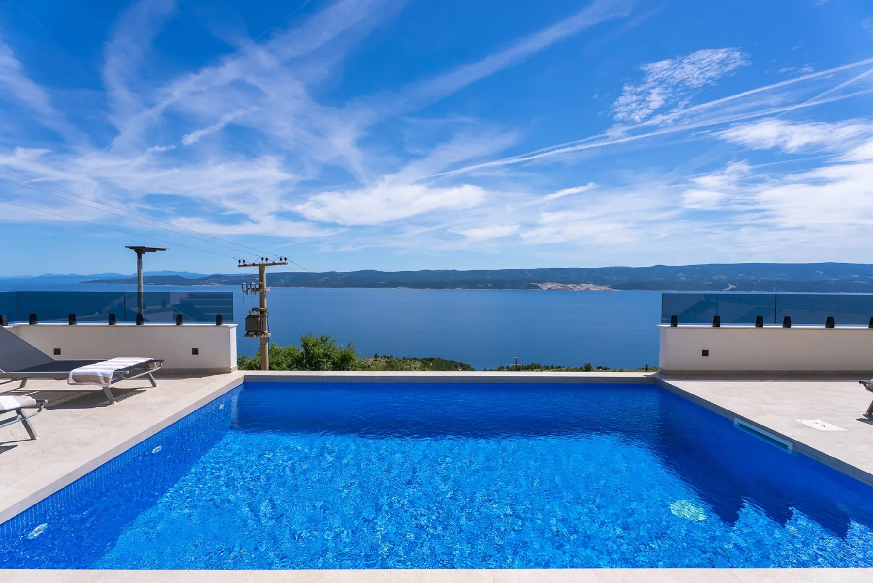 An outdoor swimming pool overlooks a calm blue bay, with loungers on the surrounding stone terrace.