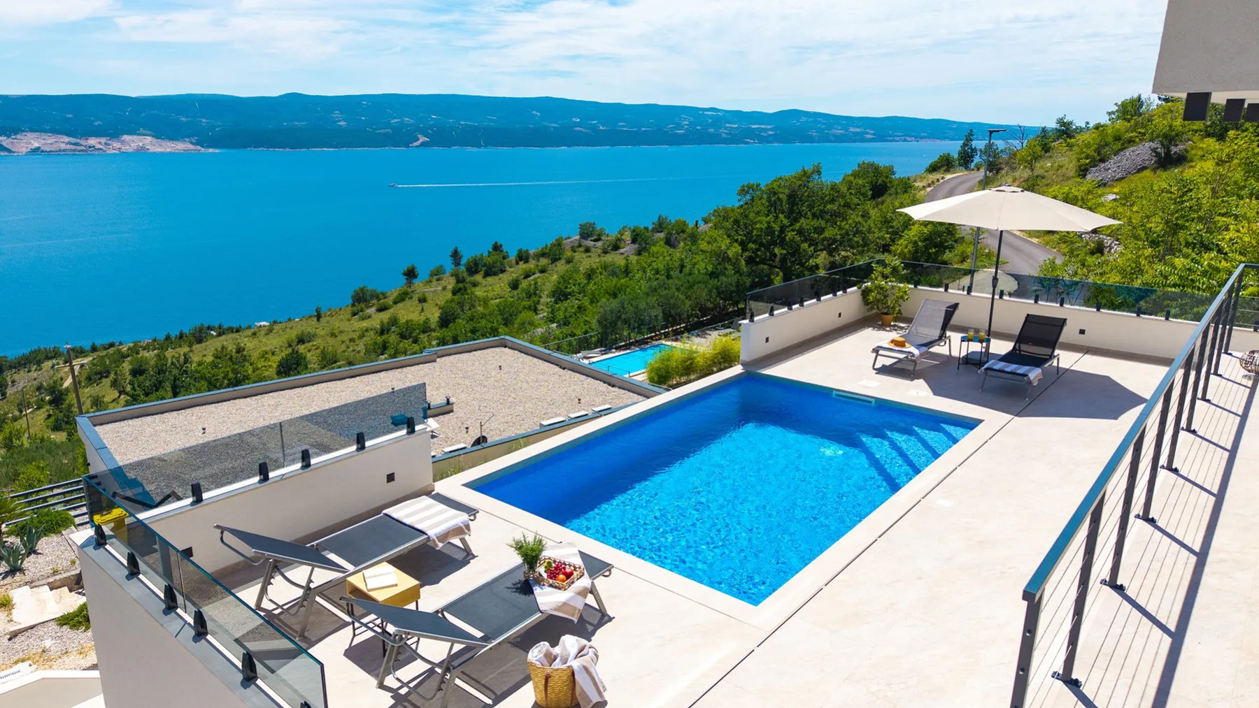 A rectangular swimming pool sits on a sunny terrace overlooking a turquoise bay and green hills.