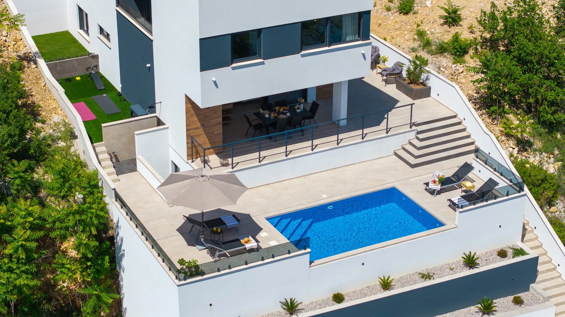 A modern house overlooks a bright blue pool, with a broad paved terrace, outdoor seating, and steps.