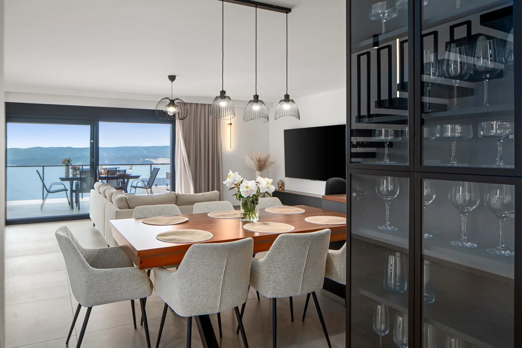 A dining table with upholstered chairs sits beside a glass-front cabinet, with a living area and balcony beyond.