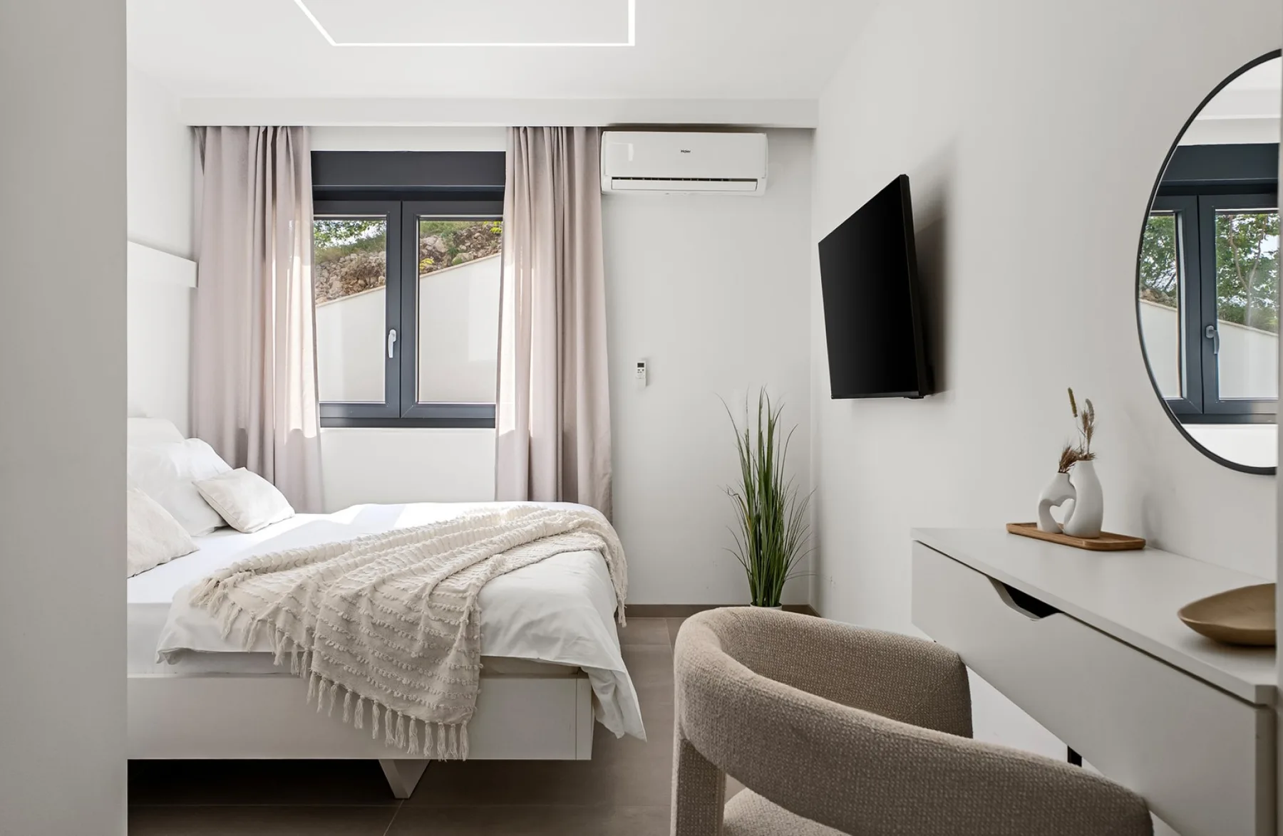 A bright bedroom has a white bed, pale curtains, a wall-mounted TV, and a chair beside a dressing table.