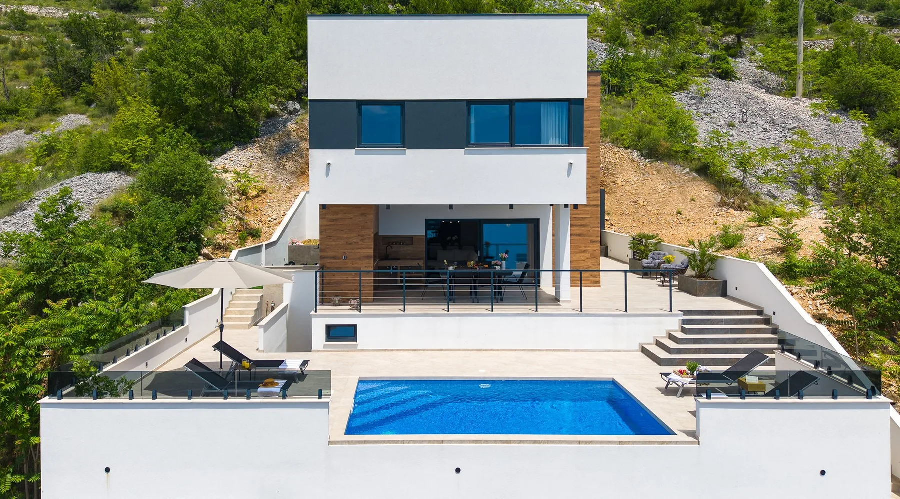 A modern white house overlooks a rectangular blue swimming pool, with lounge chairs on the surrounding terrace.
