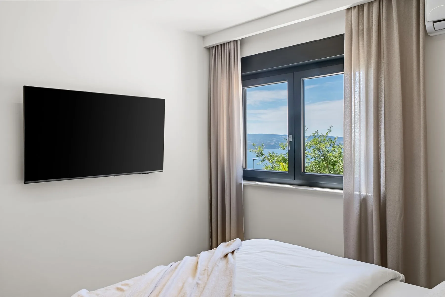 A bed sits beneath a window with beige curtains, green foliage outside, and a wall-mounted television.