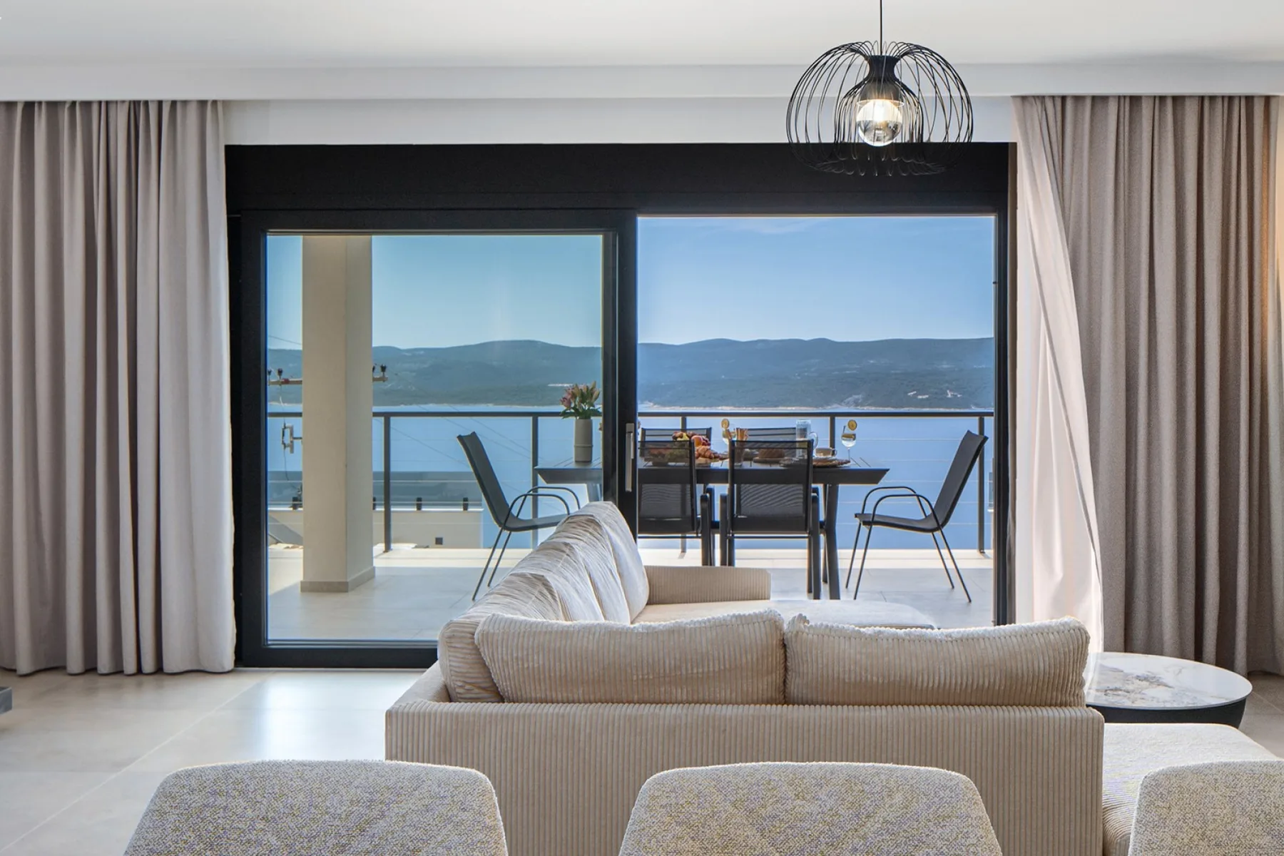 A modern living room opens onto a balcony with outdoor dining furniture and a view of the water and hills.