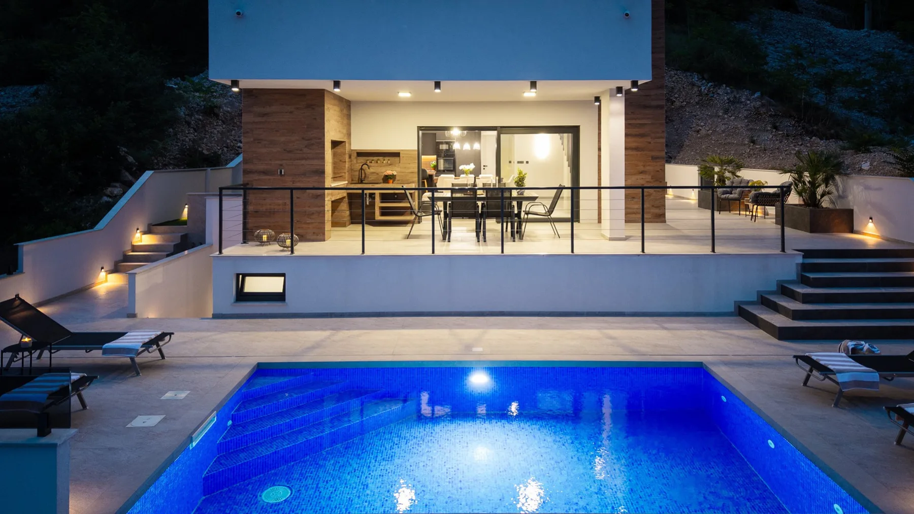 An illuminated swimming pool sits below a modern house with a glass-fronted terrace and outdoor dining table.