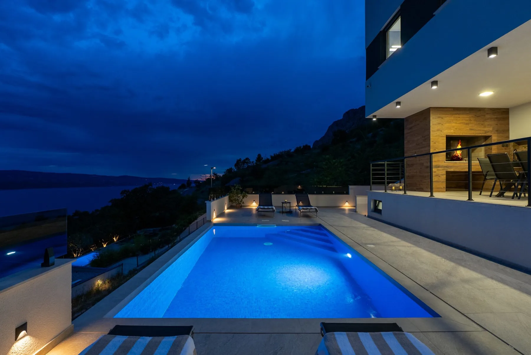 A modern house overlooks an illuminated rectangular swimming pool and distant water at dusk.