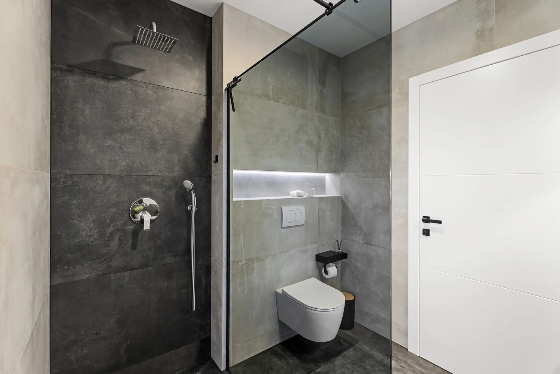 A modern tiled bathroom has a glass walk-in shower, wall-mounted toilet, and white door.