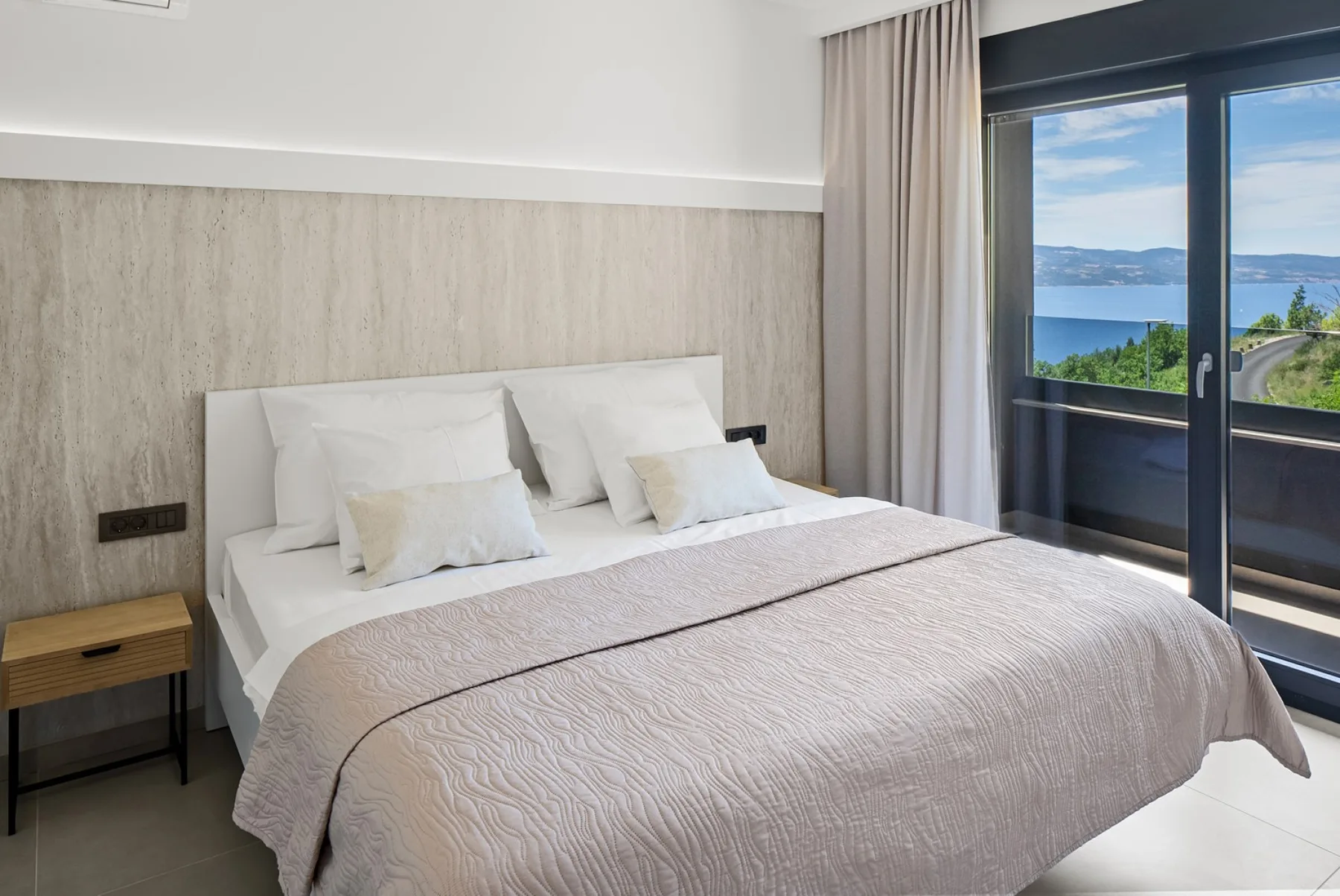 A neatly made bed with white pillows sits beside large glass doors overlooking the water.