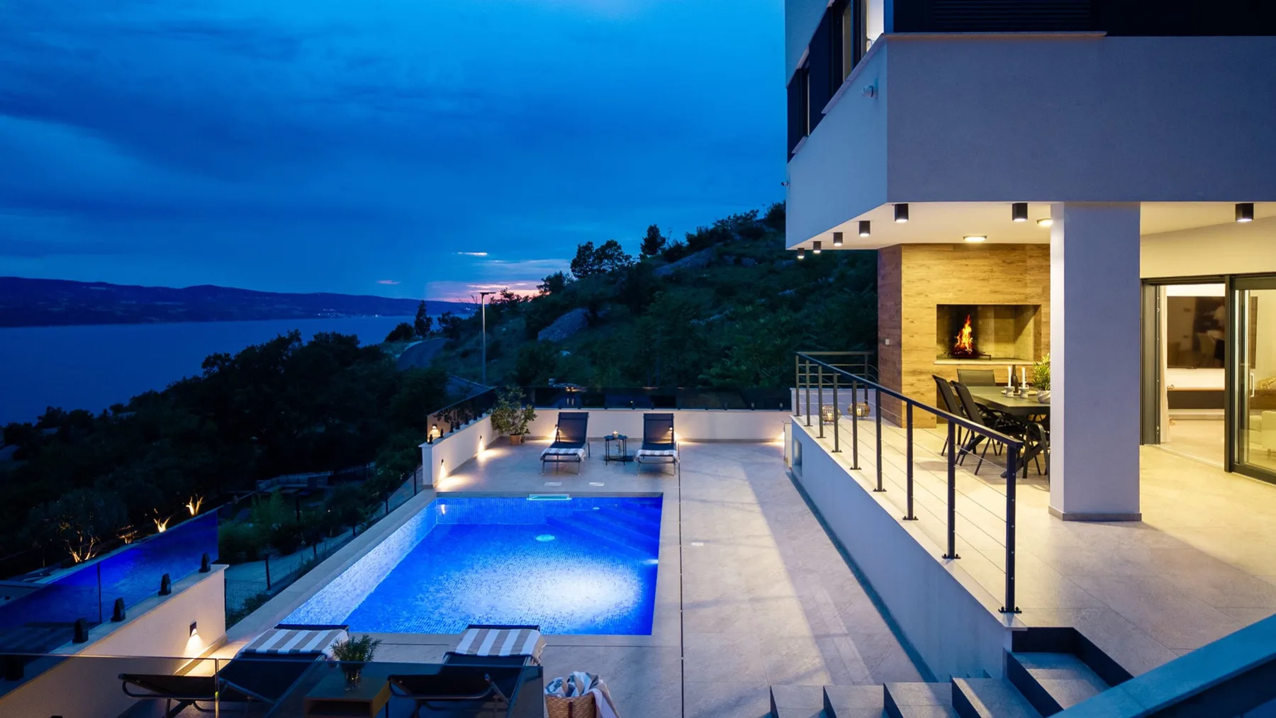 A modern house opens onto a lit swimming pool and terrace overlooking the water at dusk.