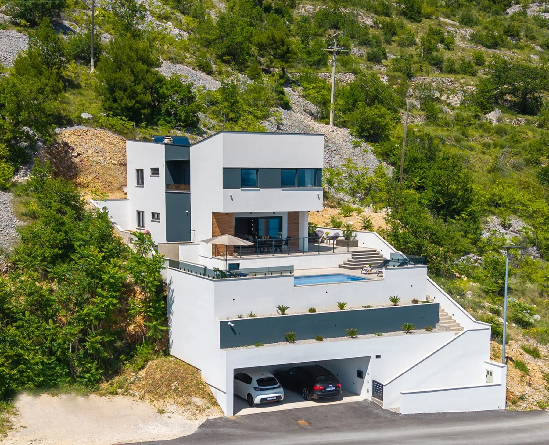 A modern white hillside house has tiered terraces, a small pool, and a two-car garage.