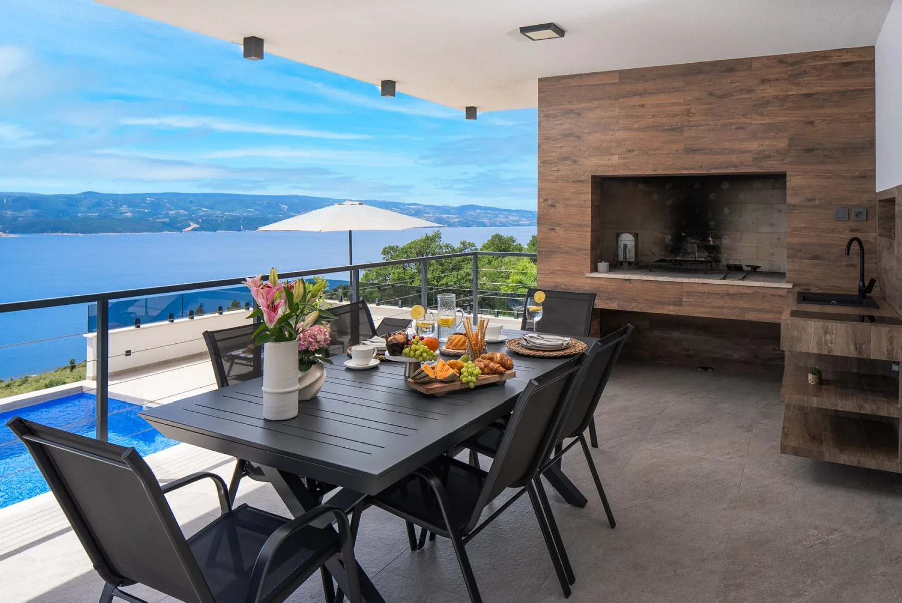 An outdoor dining table is set beside a pool, with a wood-clad barbecue area and sea views.
