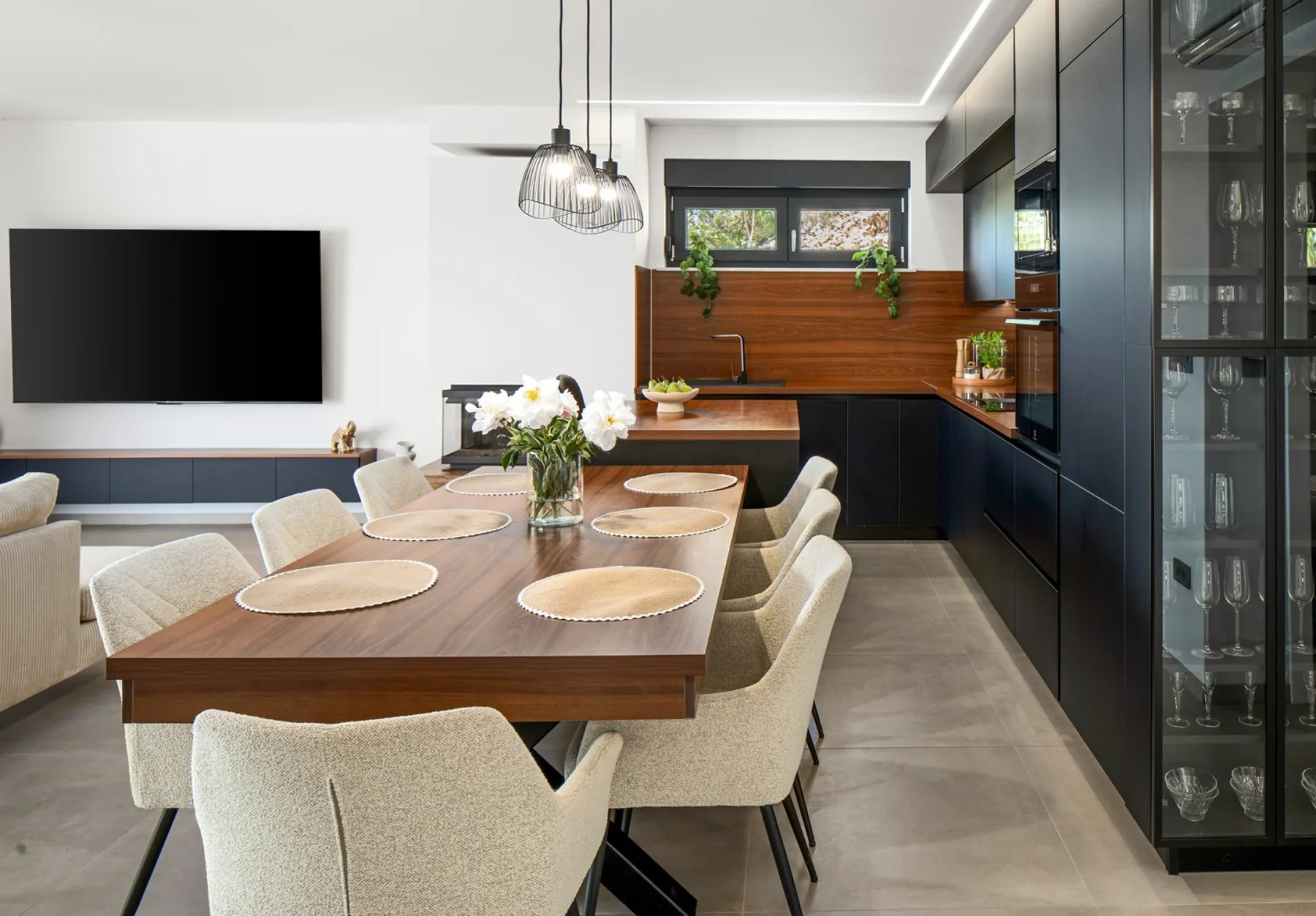 A modern dining area features a long wooden table, upholstered chairs, pendant lights, and dark kitchen cabinets.
