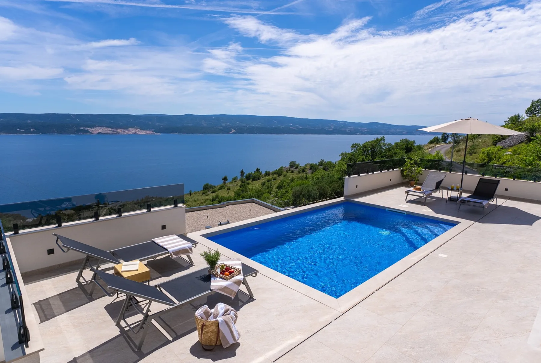 A sunlit terrace features a blue swimming pool, lounge chairs, and an umbrella overlooking a broad lake.