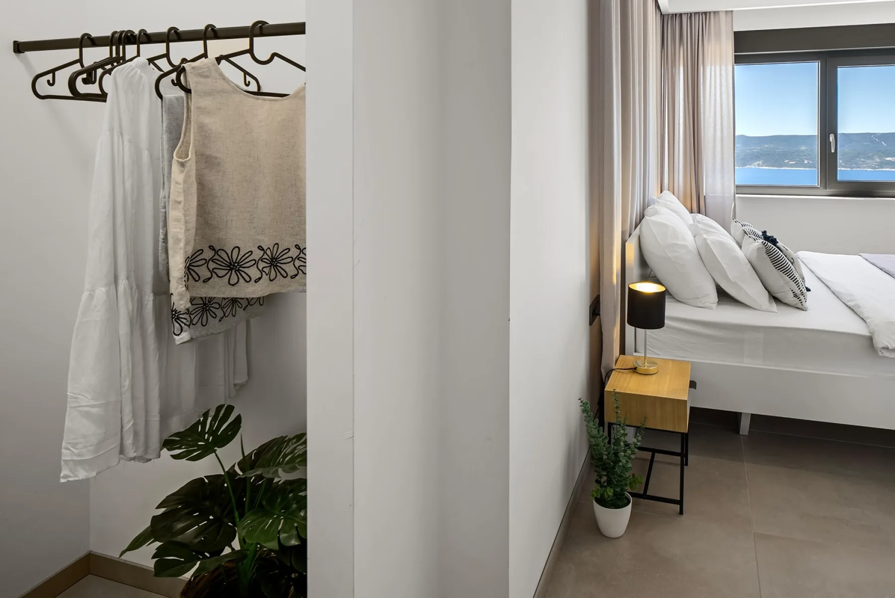 A bright bedroom has a white bed beside a window, with a clothes rack and hanging garments nearby.