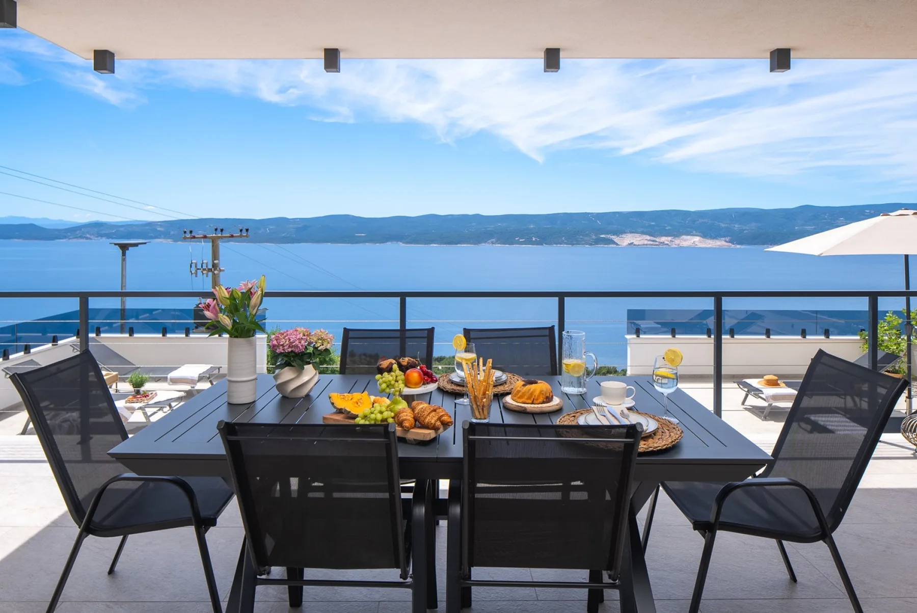 An outdoor dining table is set with food and flowers beside a railing overlooking the water and distant hills.