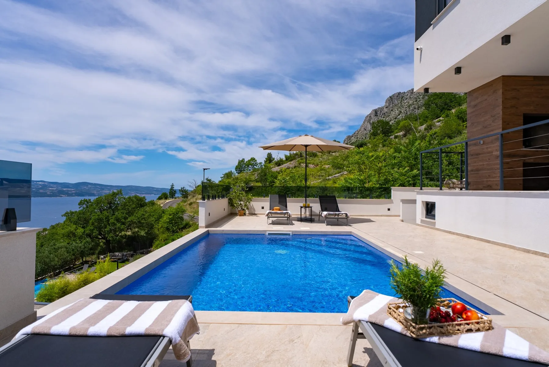 A blue swimming pool sits beside a modern villa, with sun loungers, an umbrella, and a sea view.