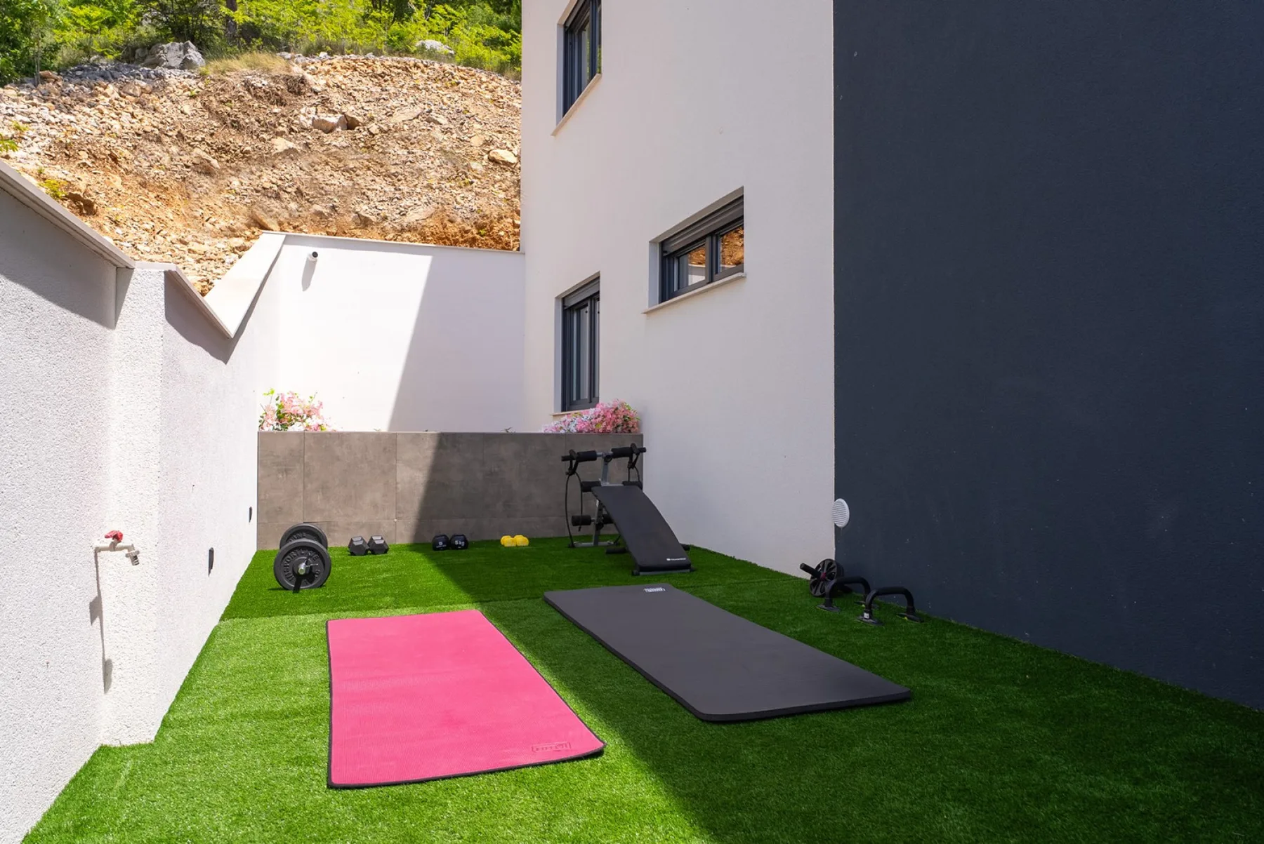 An outdoor exercise area has green turf, pink and black mats, and workout equipment beside white walls.