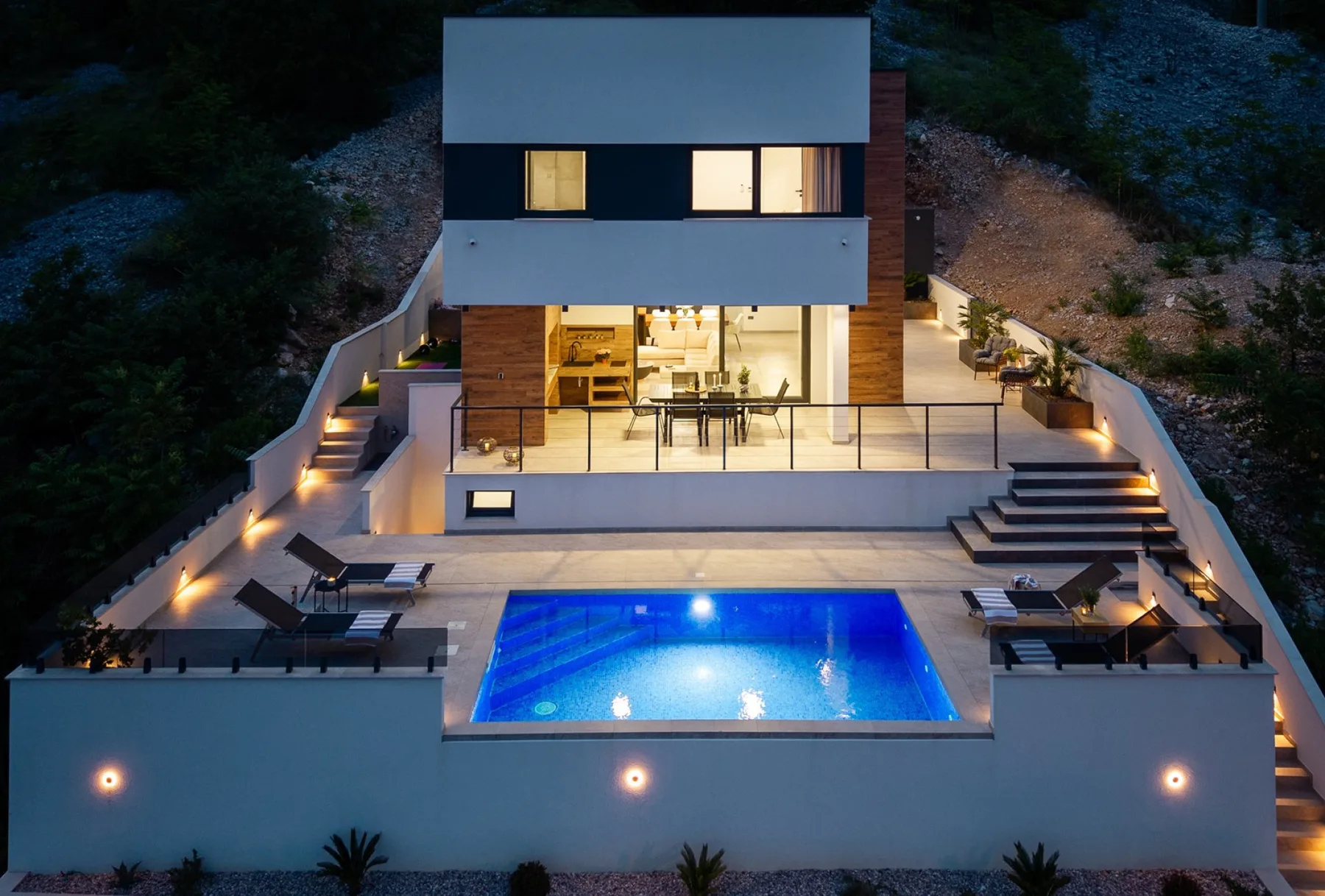 A modern hillside house overlooks a lit swimming pool, with terraces, outdoor seating, and steps.