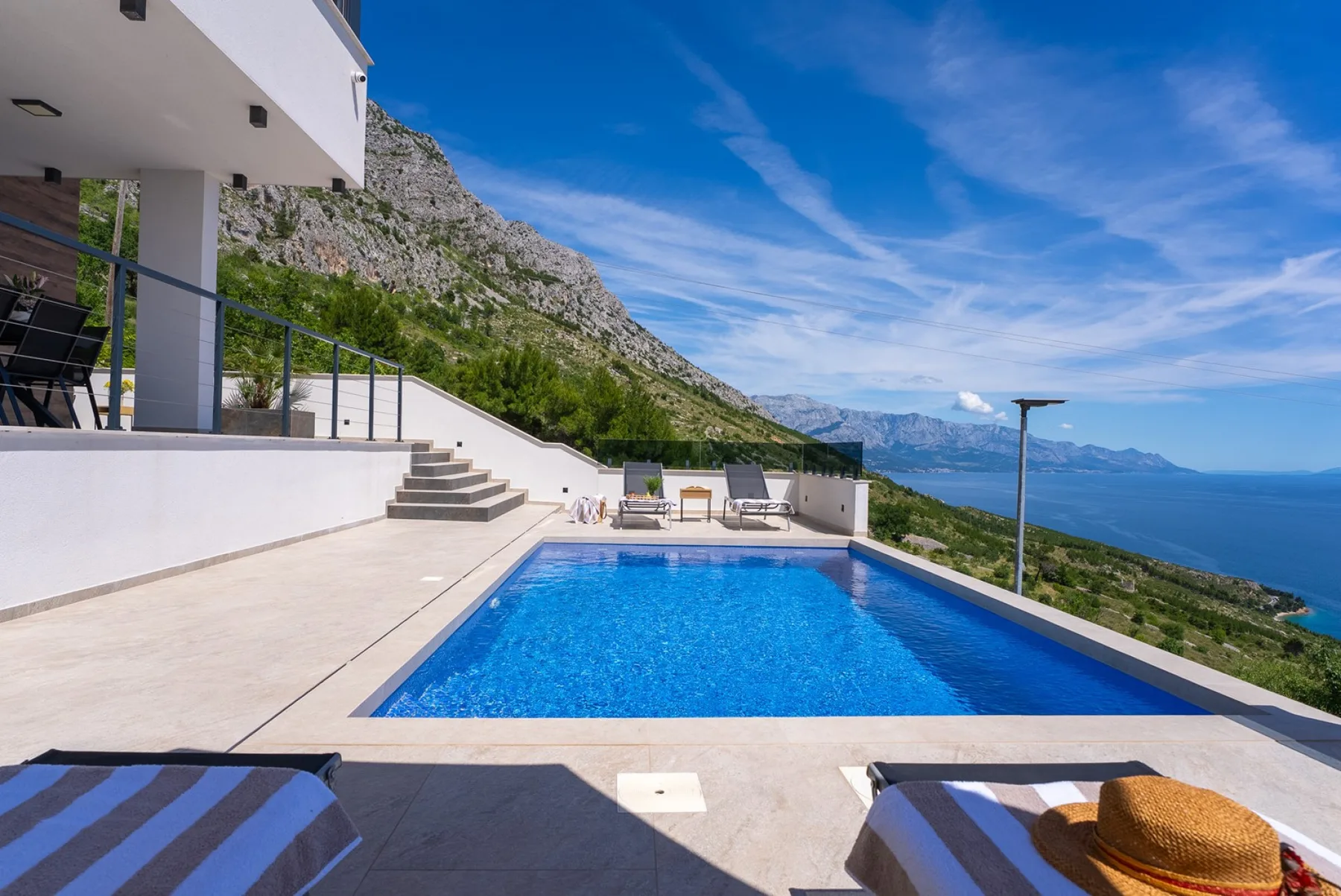 A rectangular swimming pool sits on a sunlit terrace, with loungers and a mountain-and-sea view.