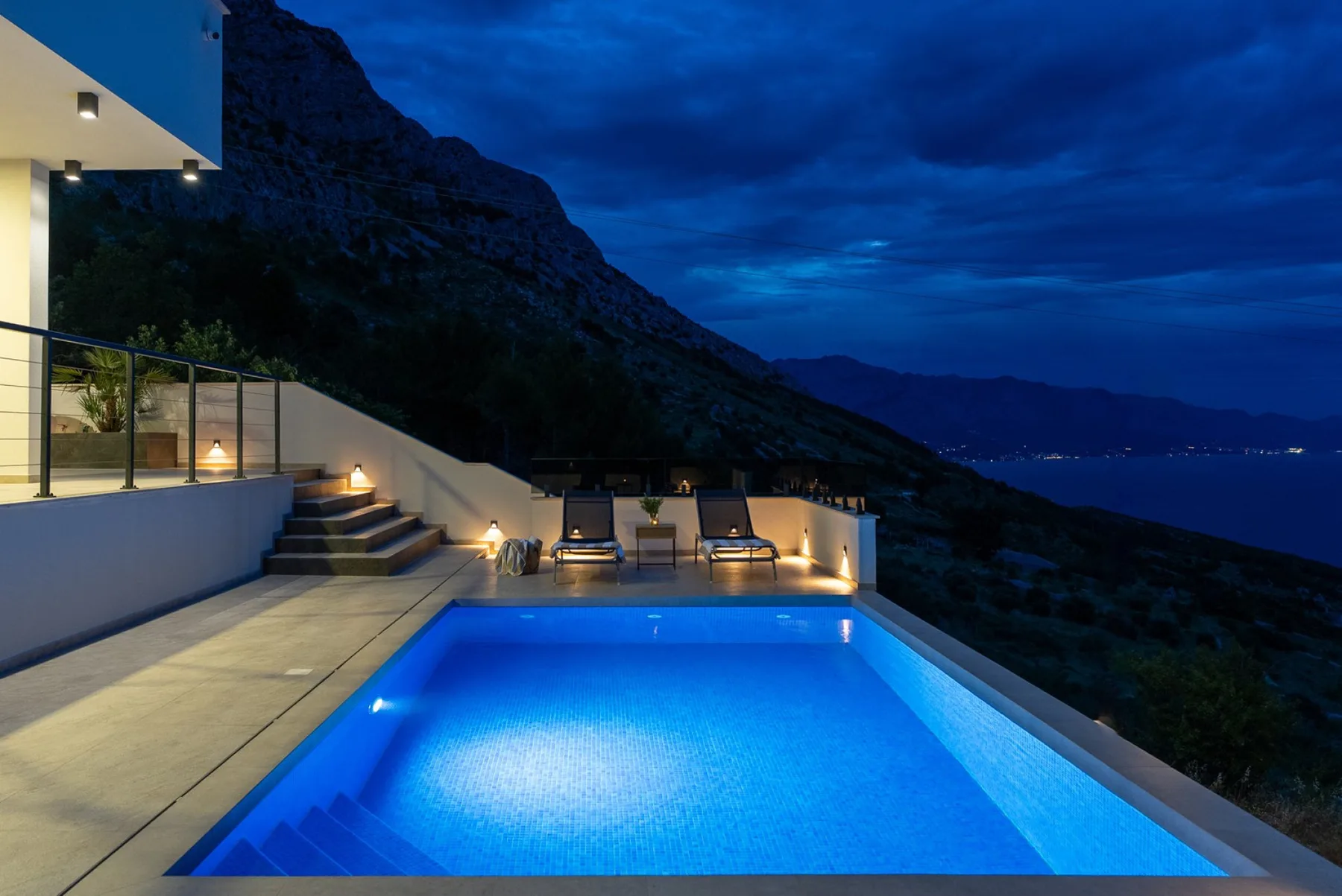 A blue-lit outdoor pool sits beside a modern terrace with steps, lounge chairs, and a mountain view at dusk.