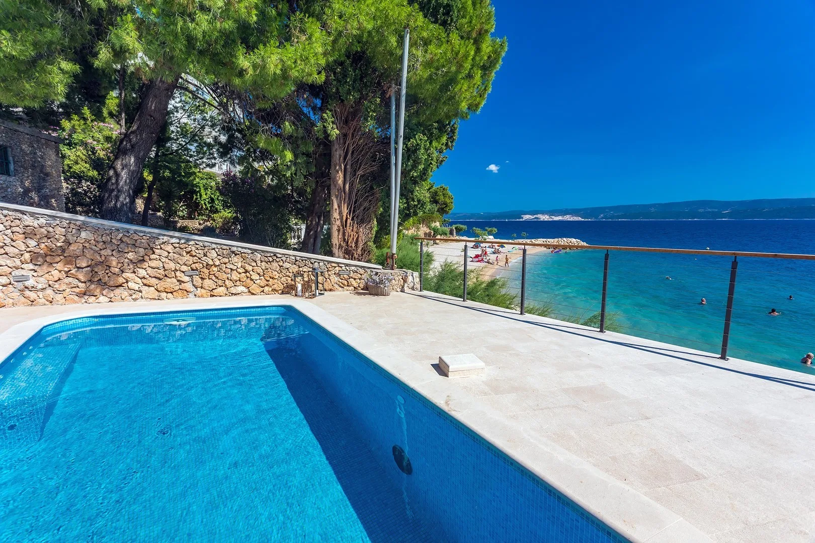 A swimming pool beside the sea, with a stone wall and trees nearby.