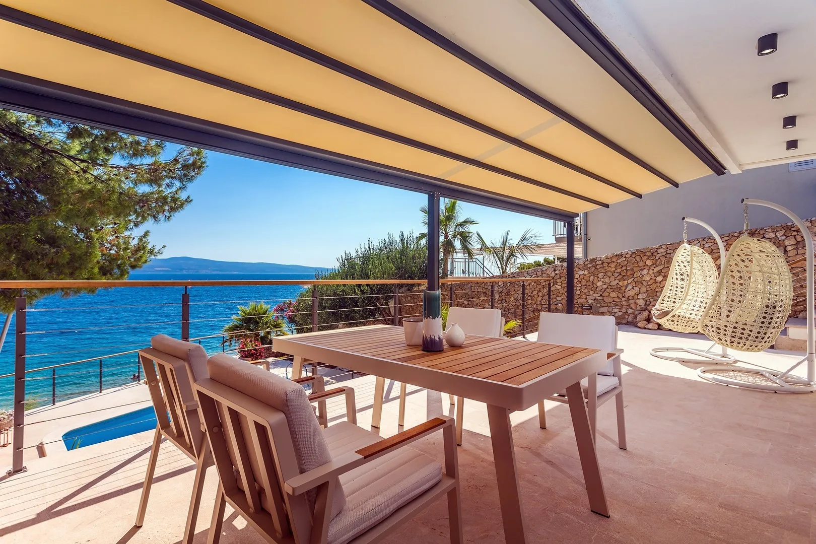 A covered seaside terrace has a wooden dining table, seating, and views of the water.