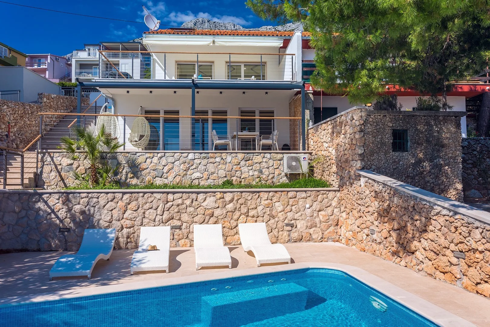 A swimming pool and loungers sit below a multi-level house with balconies.