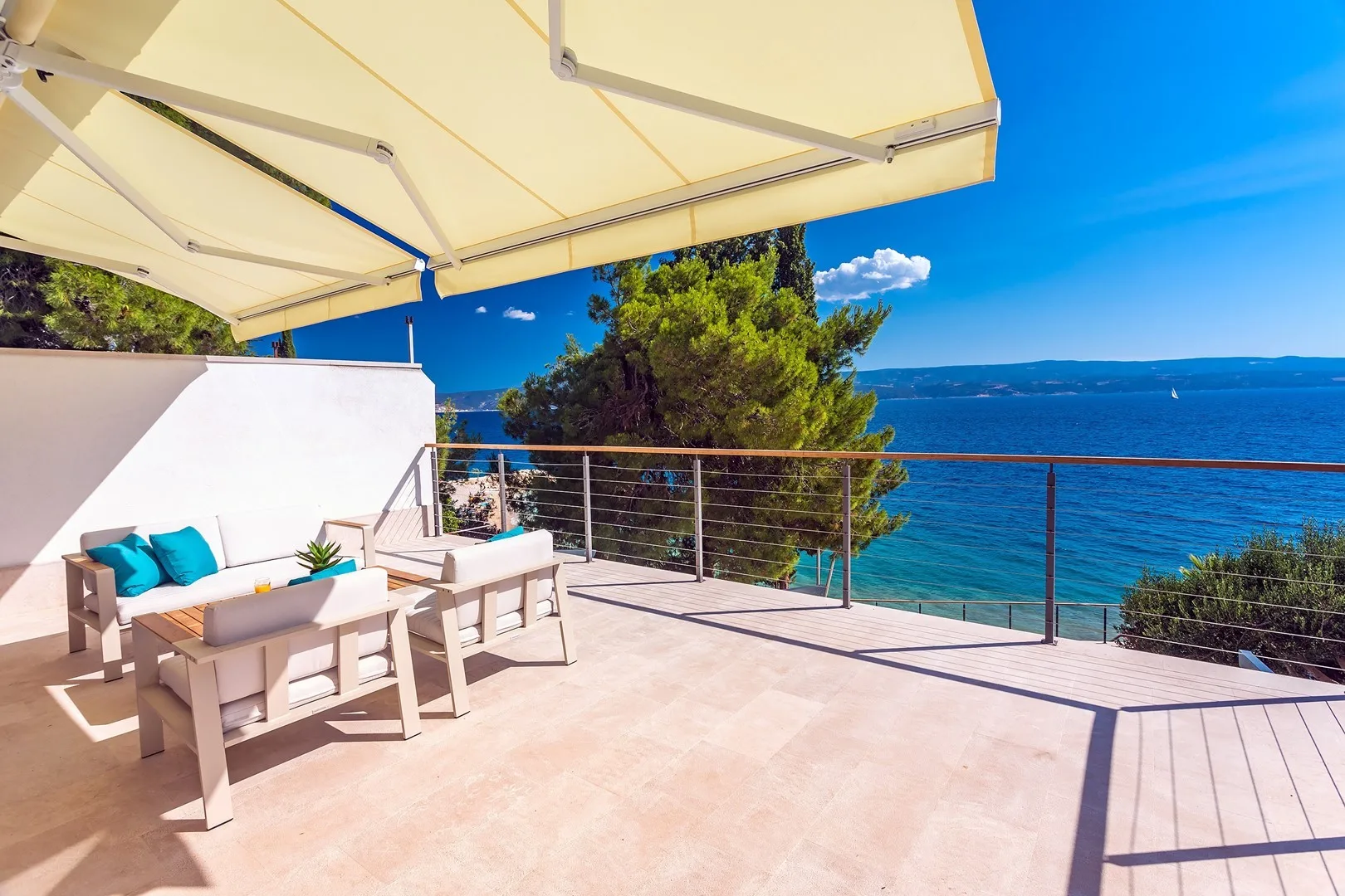 A sunlit terrace overlooks the sea, with cushioned seating beneath a large awning.