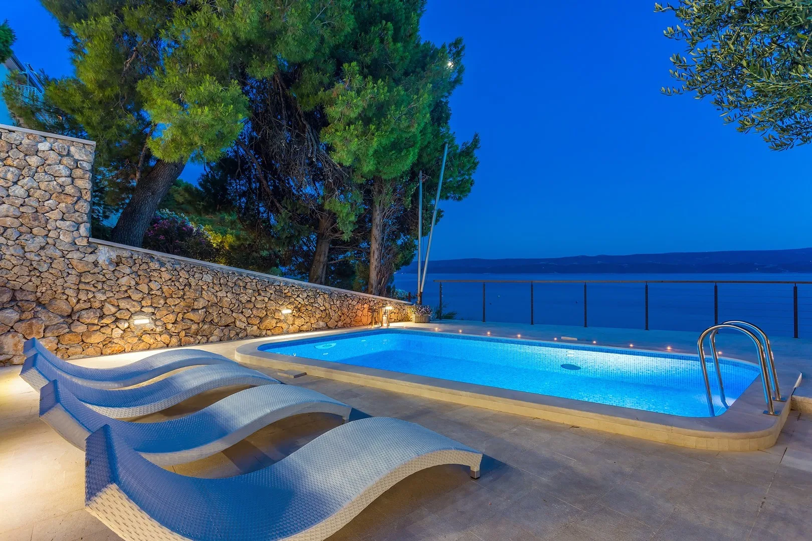 An illuminated outdoor swimming pool sits beside a stone wall and trees.