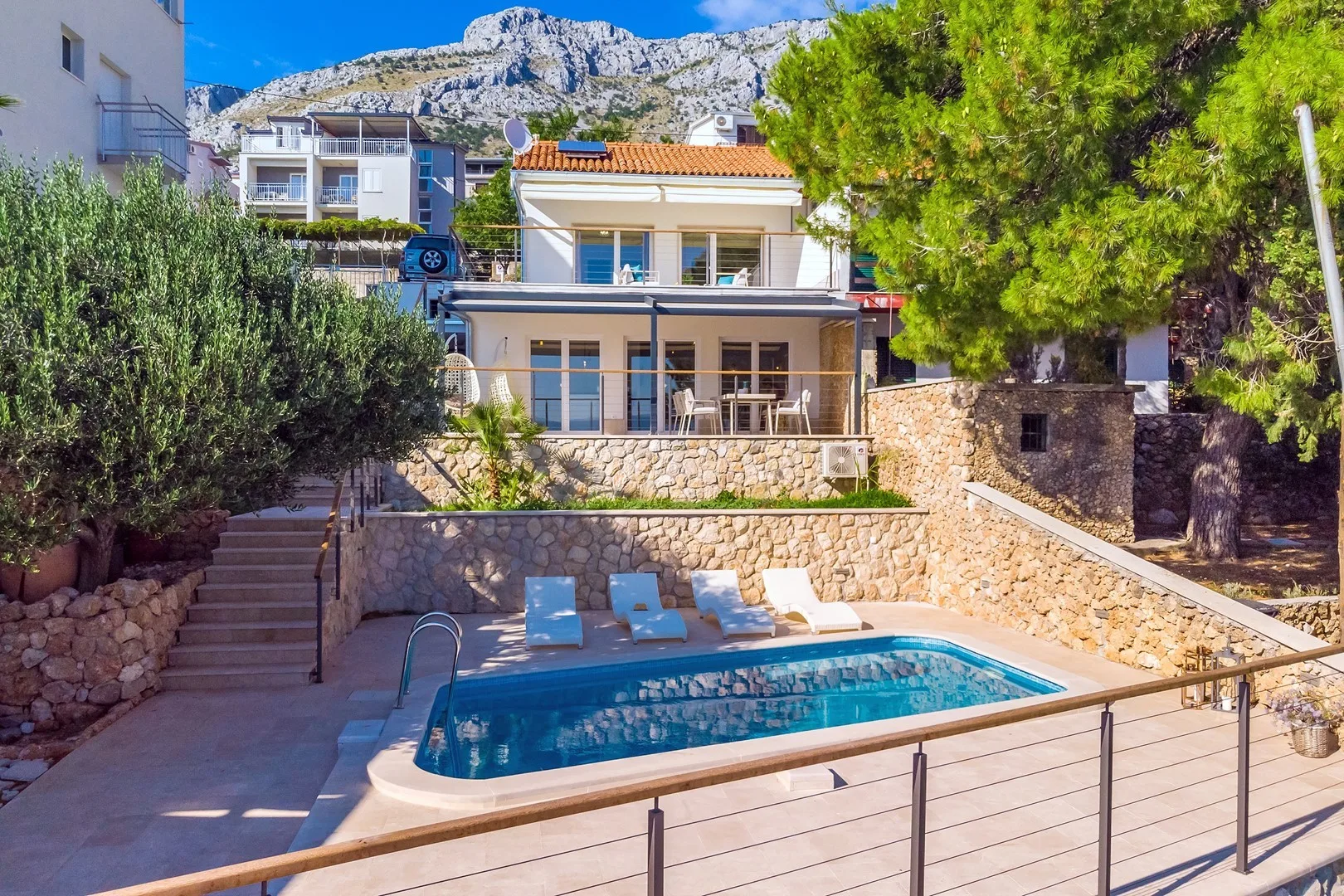 A swimming pool sits on a terrace below a multi-story house, with mountains behind.