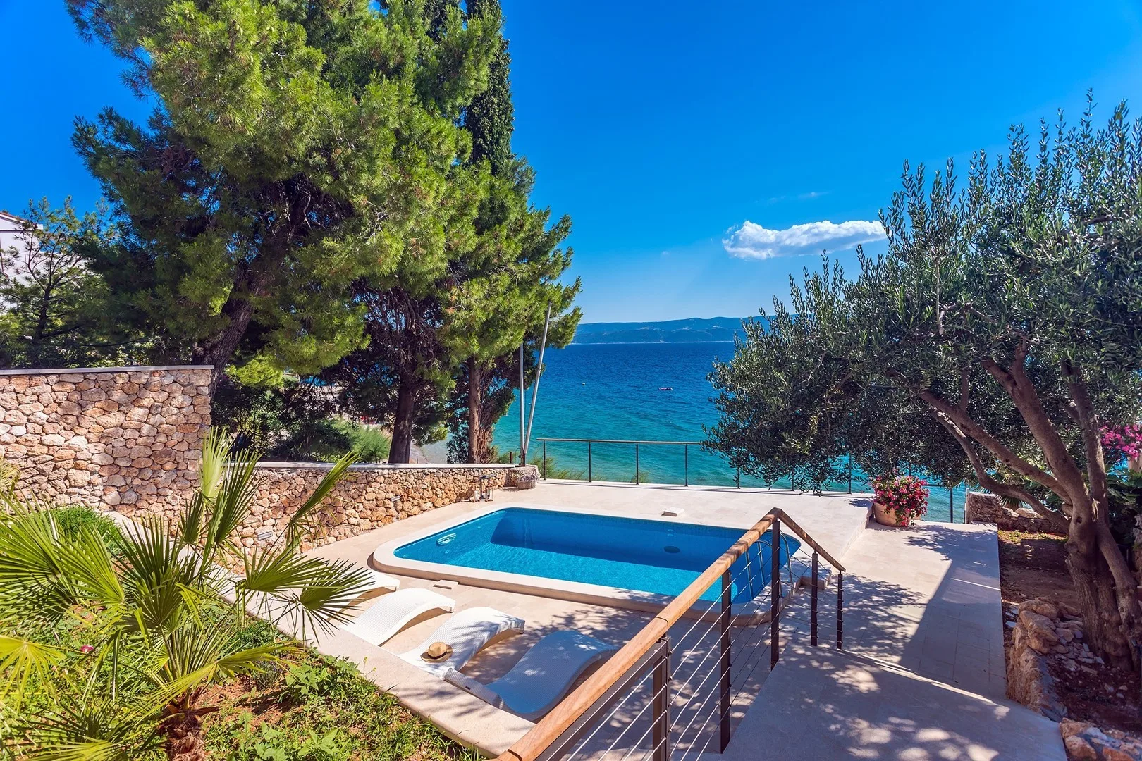 An outdoor swimming pool overlooks the sea, surrounded by trees and stone walls.