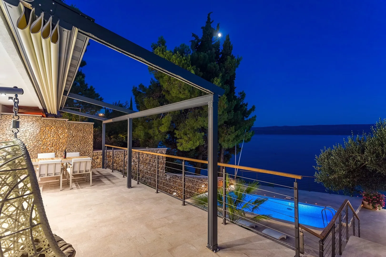 A covered outdoor terrace overlooks a lit swimming pool and the sea at dusk.