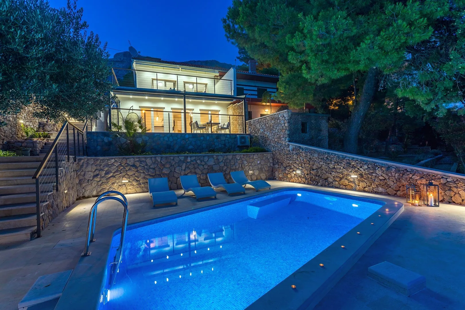 A swimming pool at dusk sits below a modern house and stone terrace.