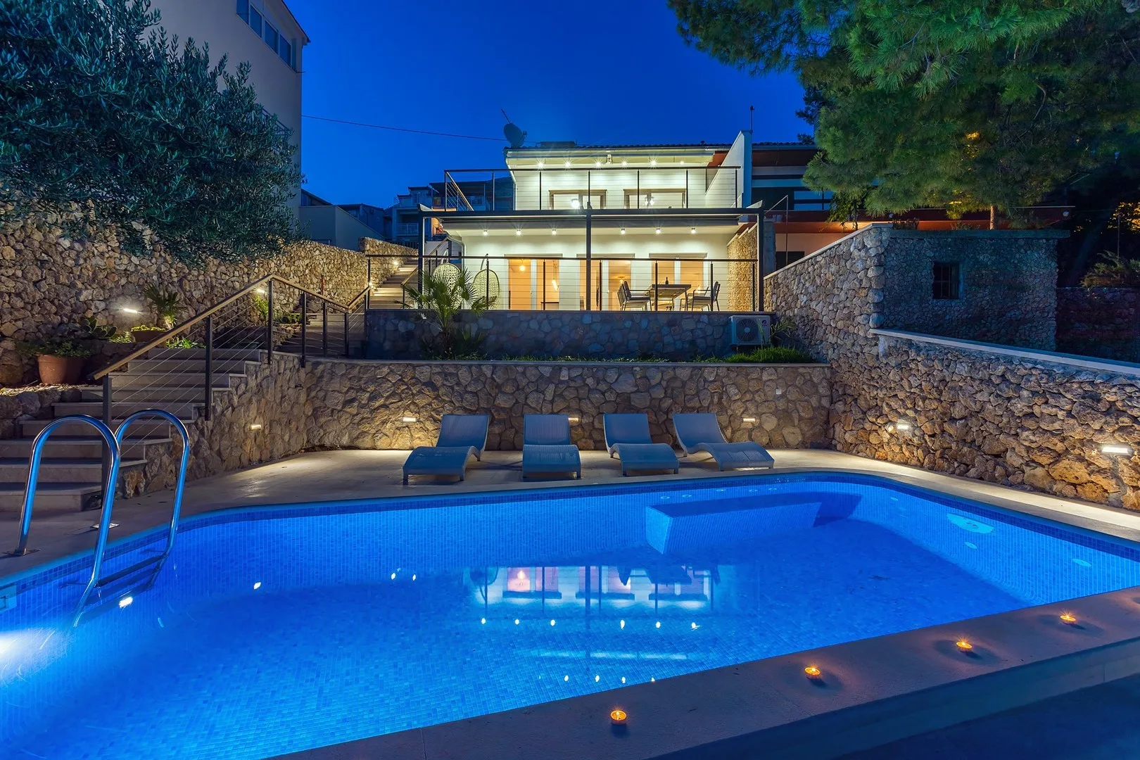 A swimming pool at dusk sits below a brightly lit house and stone terrace.
